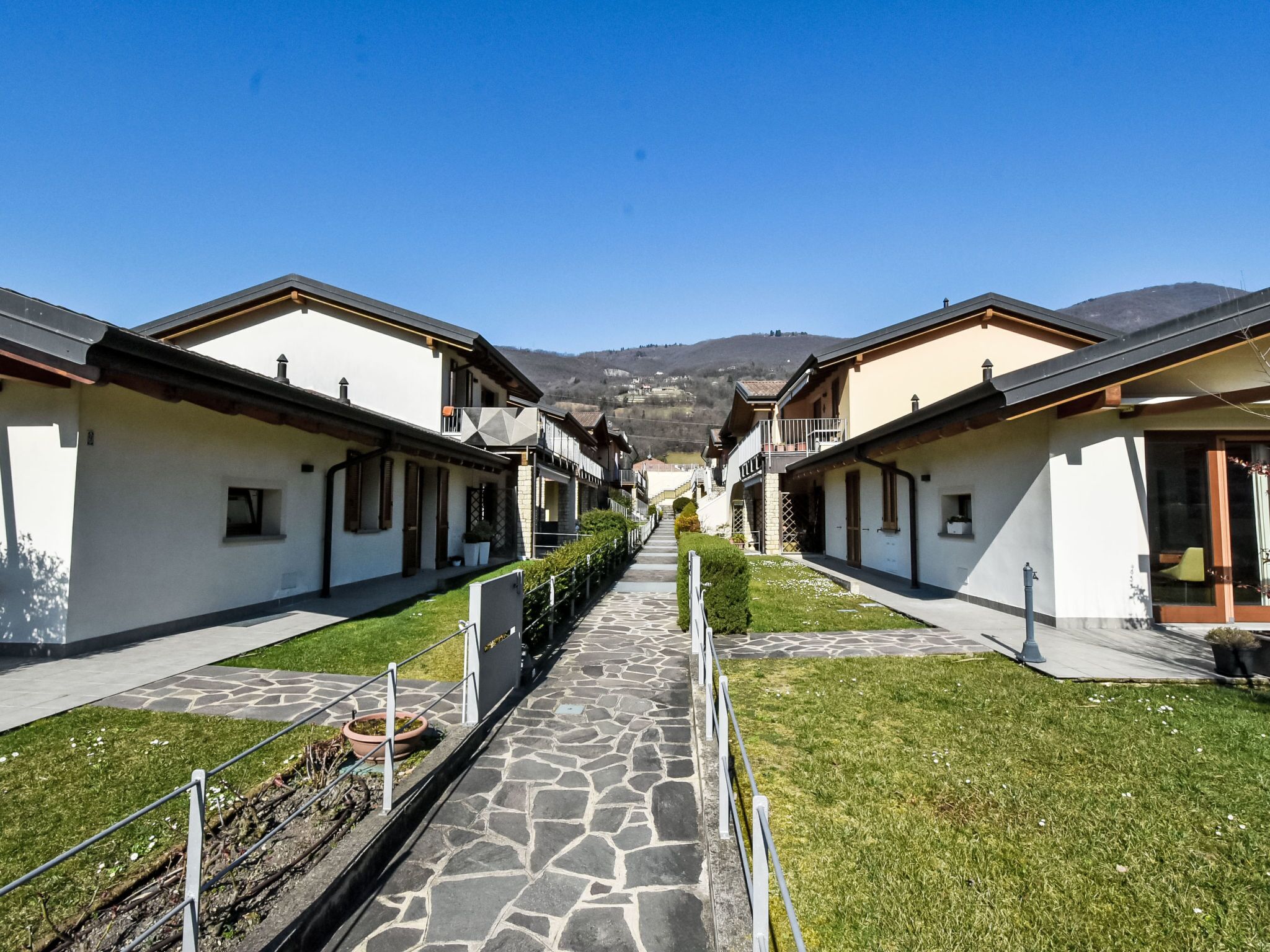 A row of white buildings with stone pathways and green lawns, leading towards a mountain.