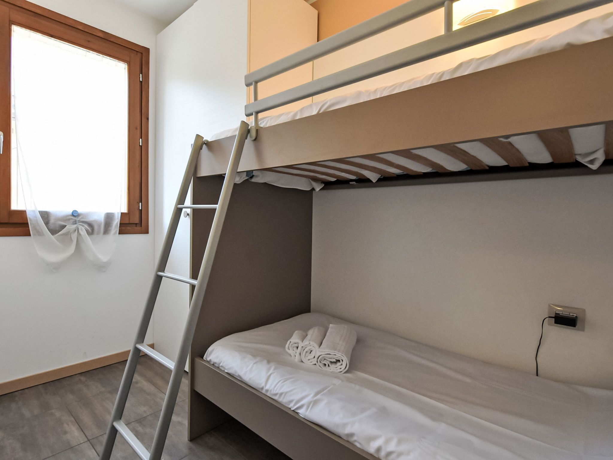 A bunk bed with a ladder, a window with a sheer curtain, and a wall socket.
