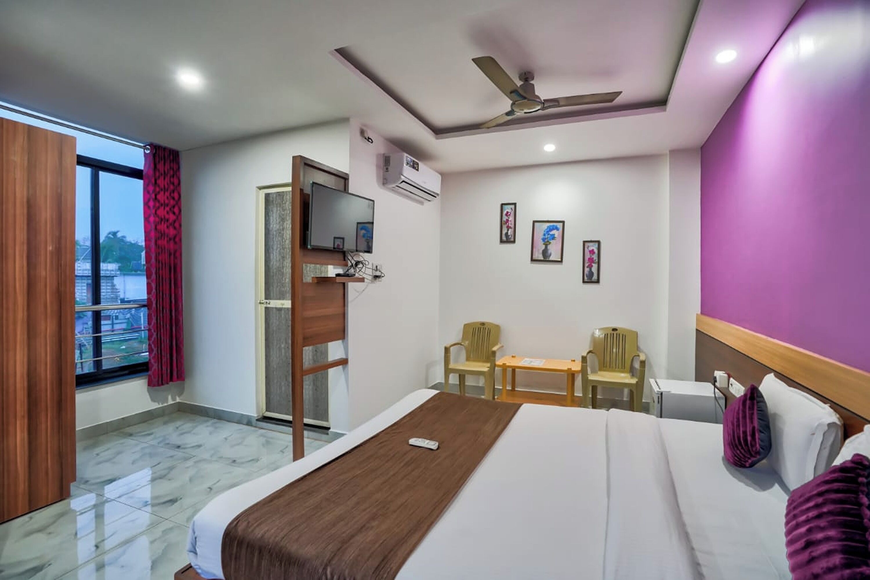 Habitación de hotel con cama, escritorio, dos sillas, televisor, aire acondicionado y ventana con cortinas.