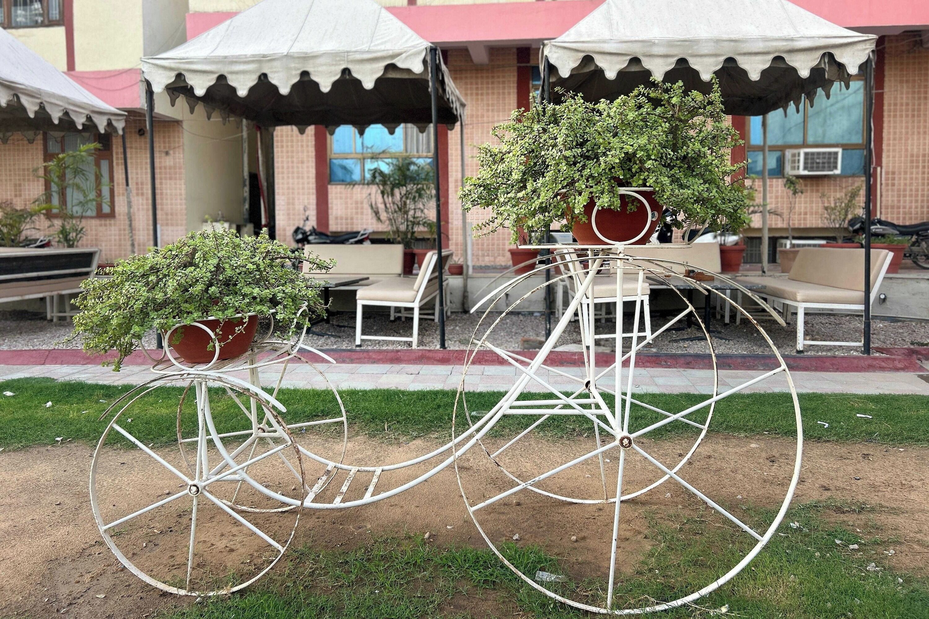 Un vélo orné de plantes en pot sur ses roues, placé dans un espace extérieur aménagé avec des chaises et des tables, et un bâtiment à l’arrière-plan dont la façade est rouge et beige.
