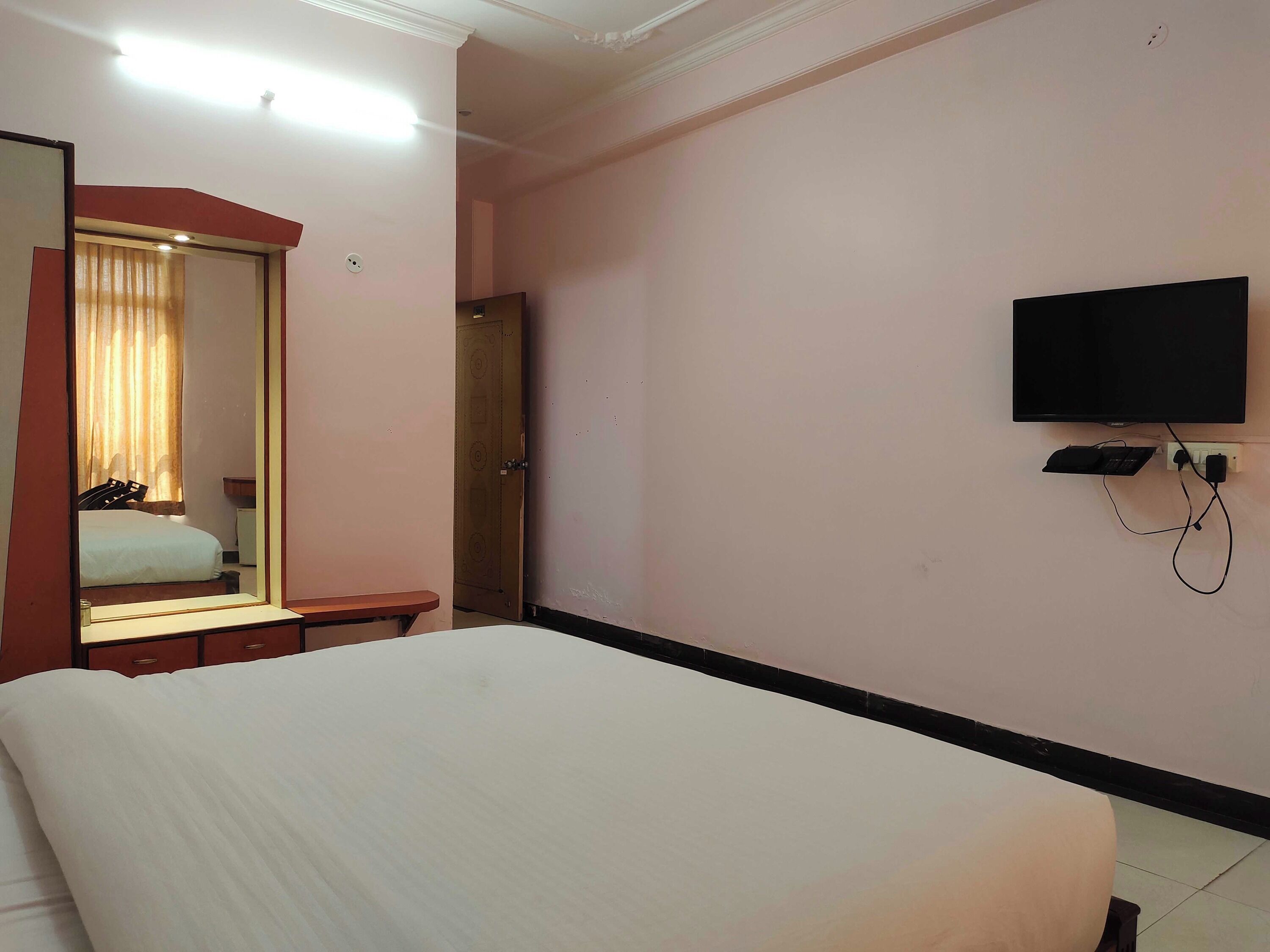 A hotel room with a bed, a bedside table, a mirror, a television mounted on the wall, and a door leading to another room.
