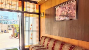 Una habitación de hotel con un sofá, una mesa de vidrio, un ventanal grande con celosías decorativas y un cuadro en la pared.