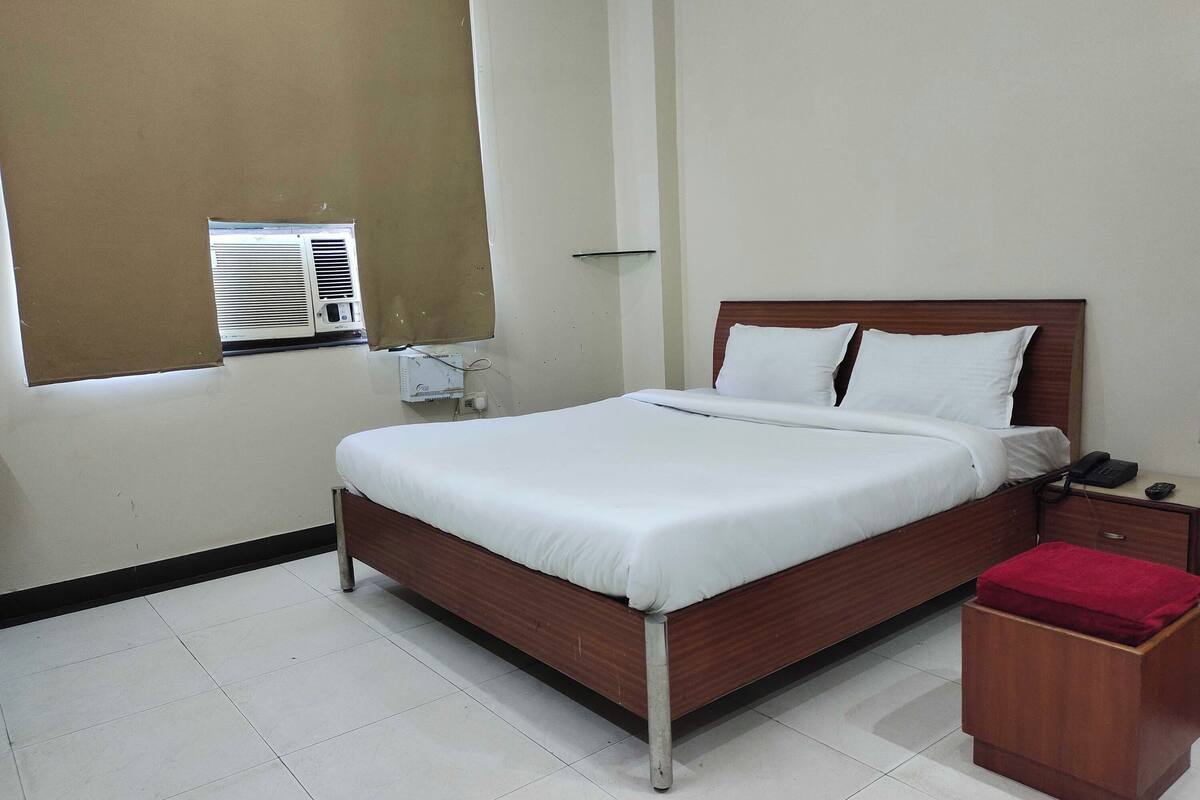 Una cama bien hecha con sábanas blancas, cabecera de madera y mesita de noche con un teléfono y un paño rojo.