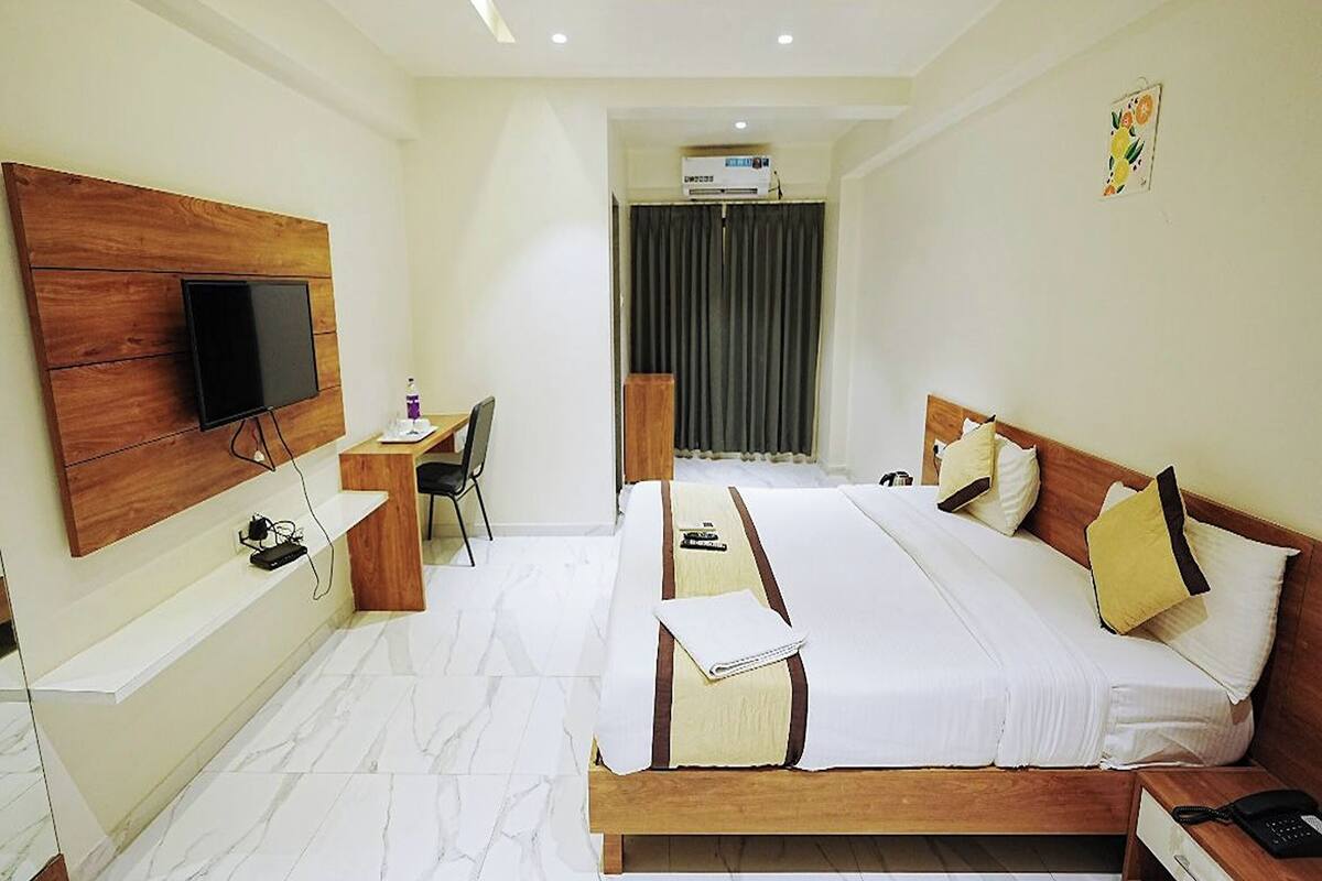 A hotel room with a wooden bed, a desk with a chair, a flat-screen TV mounted on the wall, and an air conditioning unit.