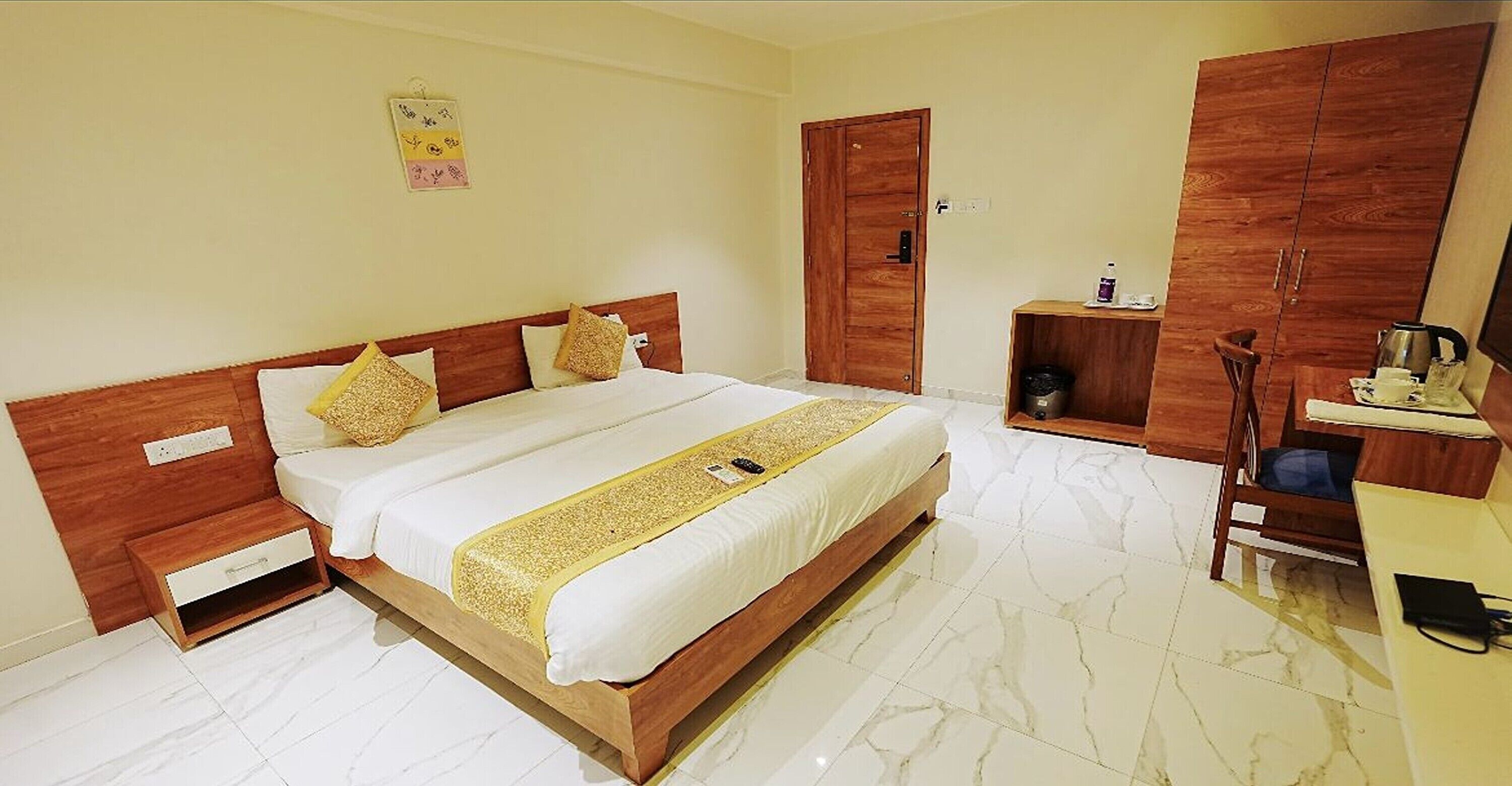 A hotel room with a large bed, wooden headboard, a bedside table, a wardrobe, and a small desk with a chair.