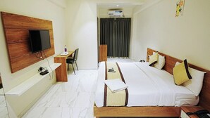 Ένα δωμάτιο ξενοδοχείου με ένα μεγάλο κρεβάτι, ένα γραφείο με καρέκλα, μια τηλεόραση τοποθετημένη στον τοίχο και μια μονάδα κλιματισμού στο βάθος.