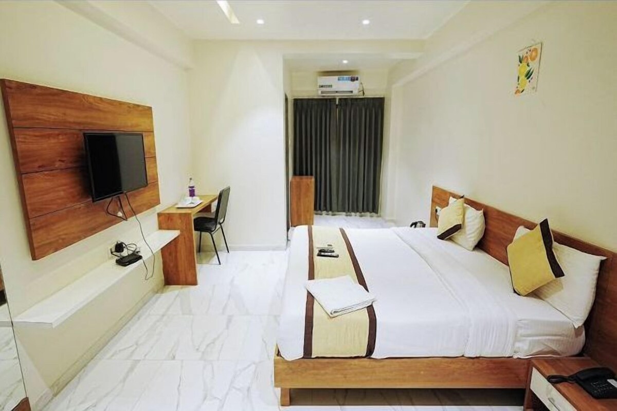 A hotel room with a wooden headboard, a flat-screen TV mounted on the wall, a desk with a chair, a bed with white bedding, and a bedside table with a phone.
