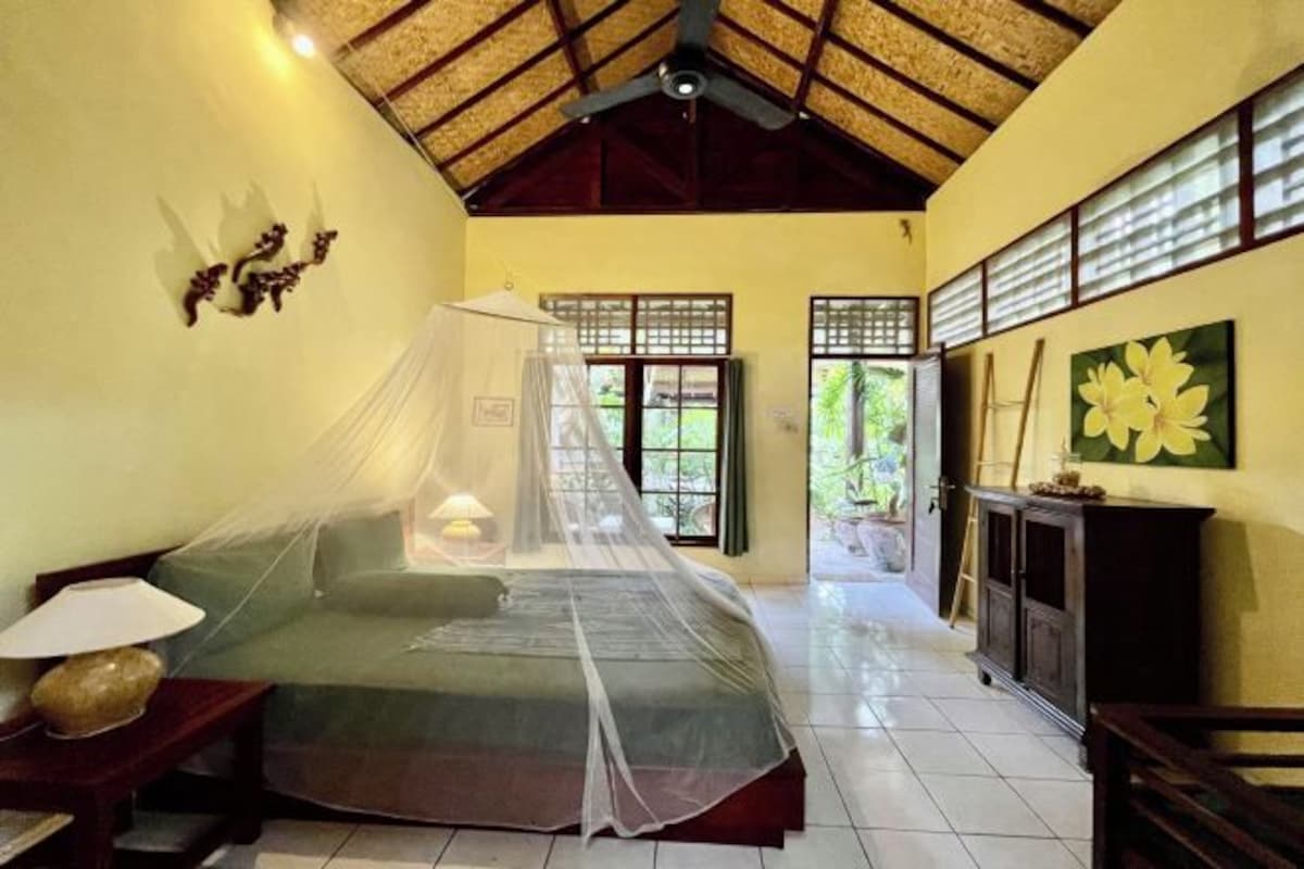 A room with a bed covered by a mosquito net, a wooden ceiling, a window with a view of greenery, and a cabinet with a flower painting.