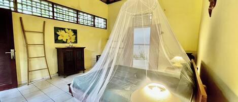 A room with a bed under a mosquito net, a wooden ladder, a lamp, and a painting on the wall.