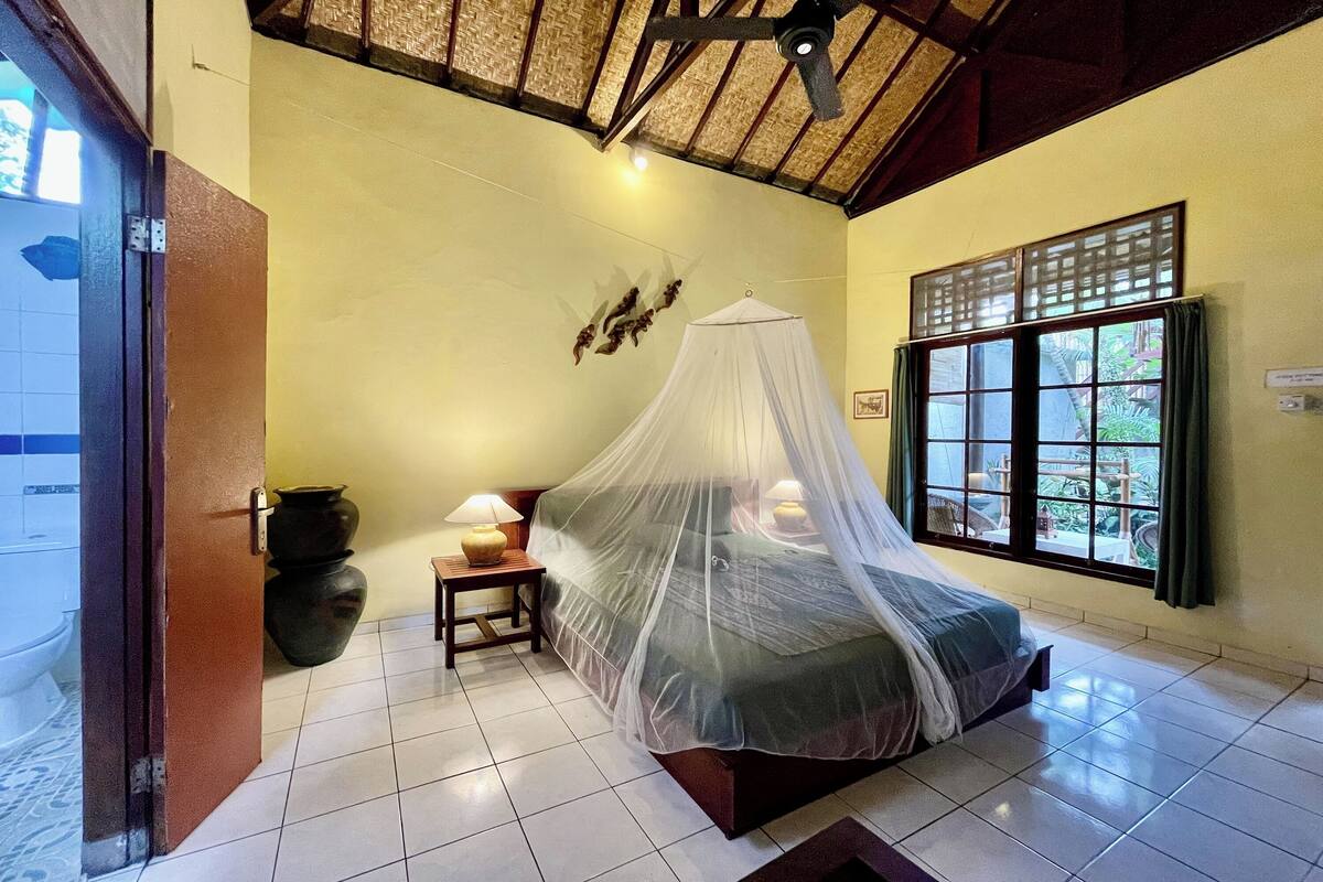A room with a bed under a mosquito net, a window with curtains, a nightstand with a lamp, and a wall-mounted decorative piece.