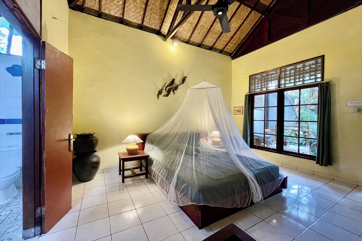 A room with a bed under a mosquito net, a window with curtains, a nightstand with a lamp, and a wall-mounted decorative piece.