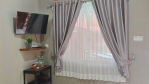 A room with a mounted TV, a shelf with decorative items, and sheer curtains on a window. - Murai Pagi Homestay (Harau)