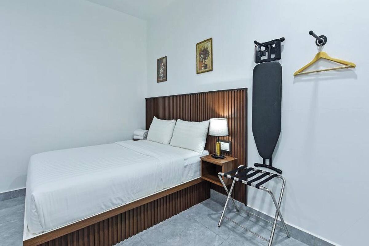 A hotel room with a bed, a nightstand, a lamp, a towel, a coat rack, and a picture frame on the wall.