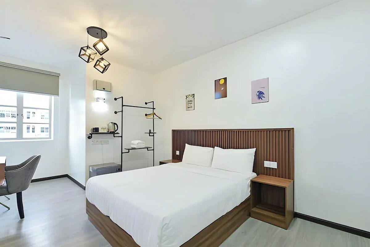 A modern hotel room with a large bed, a desk, and a chair. There is a window providing natural light, and the room has a clean, minimalist design.