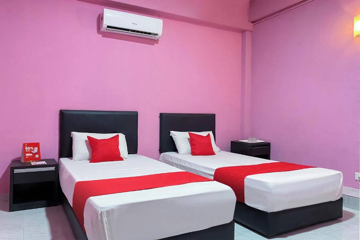Une chambre d’hôtel avec deux lits, chacun agrémenté de draps blancs et de touches de rouge, une table de chevet noire, une pancarte rouge indiquant une promotion, et un climatiseur fixé au mur.