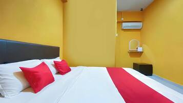 A hotel room with a bed, two red pillows, a white bedspread, a red runner, an air conditioning unit, a small bedside table with a mirror, and a black object on the floor.
