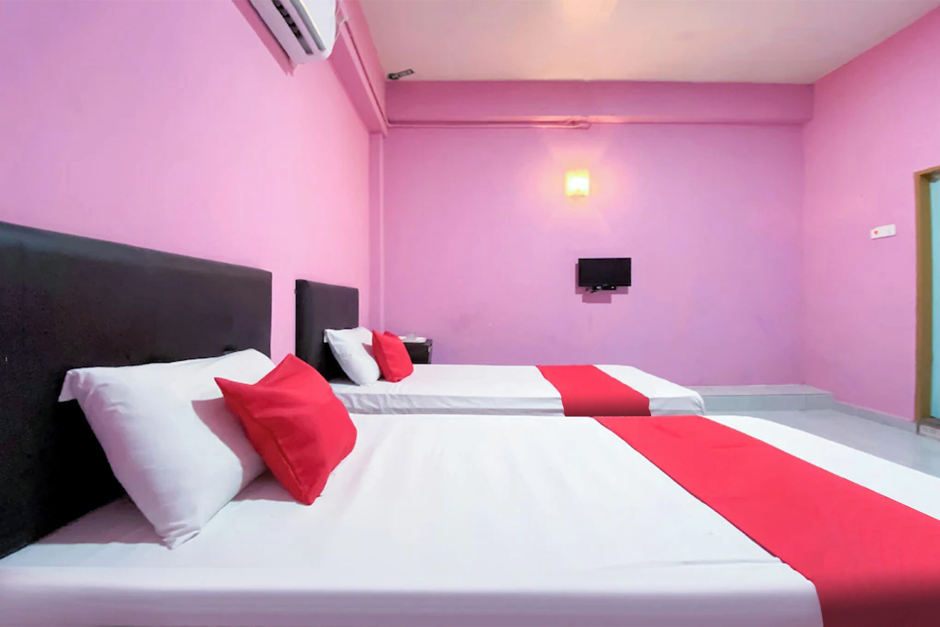 Habitación de hotel con dos camas, cada una con ropa de cama blanca y detalles rojos, paredes rosas, un televisor montado y una mesita de noche con una lámpara.