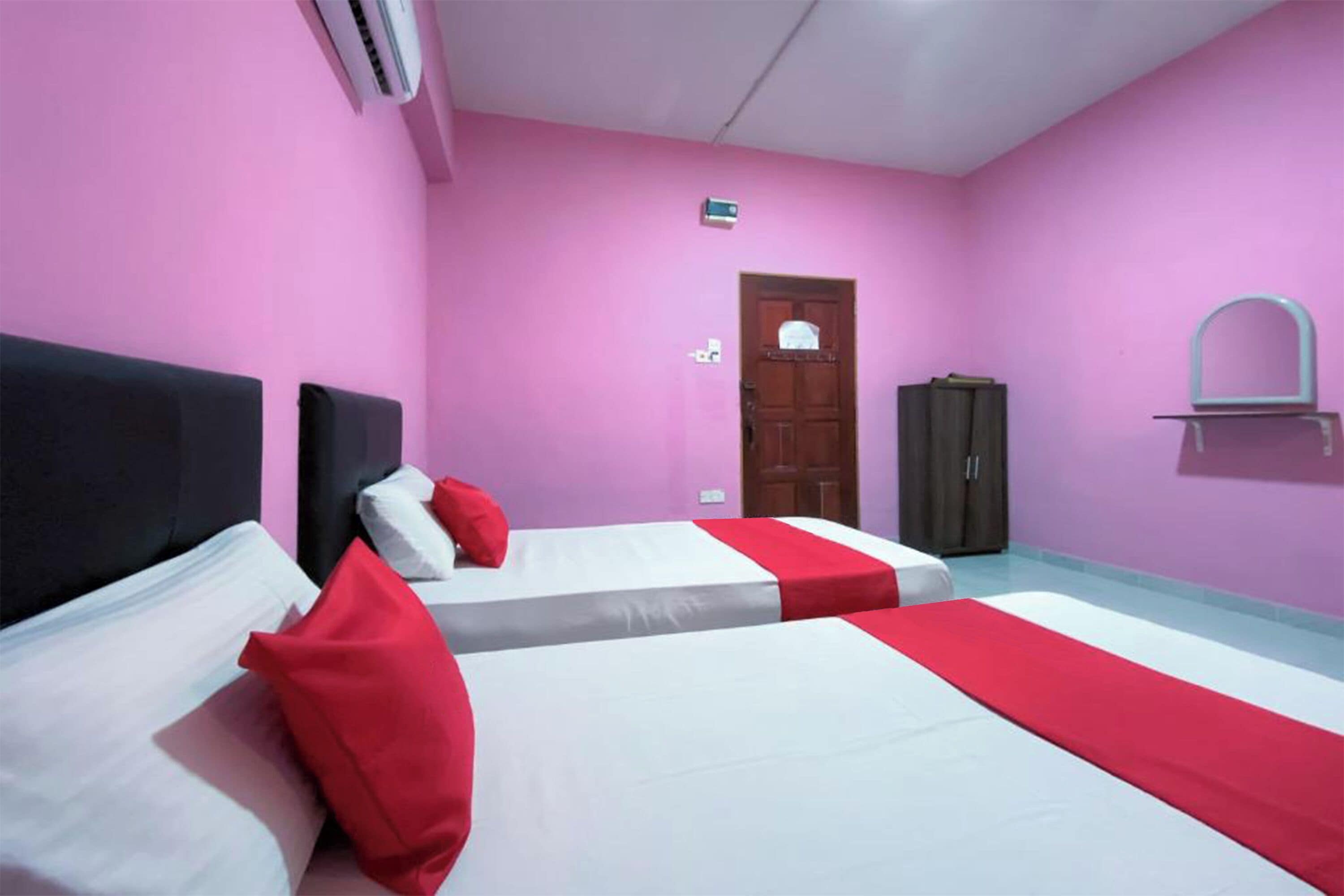 Une chambre d’hôtel avec deux lits, des murs roses, une armoire et un petit miroir accroché au mur.