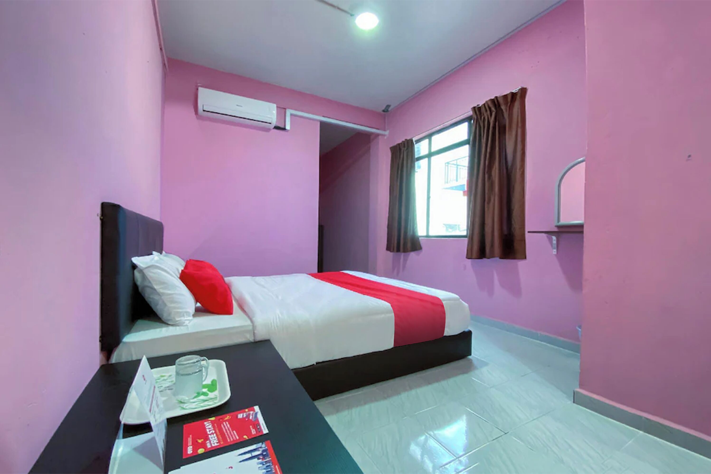 A hotel room with pink walls, a bed with white and red bedding, a table with a glass, and a window with curtains.