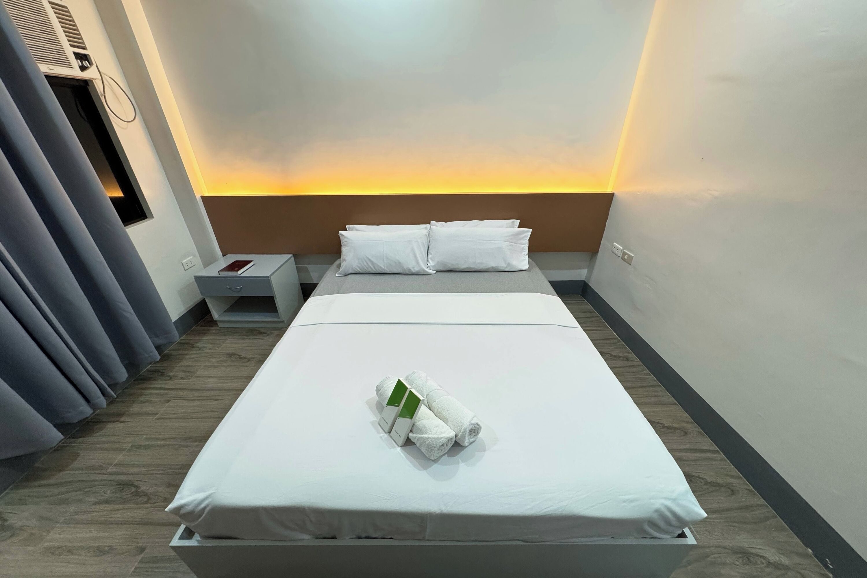 Una habitación de hotel con una cama bien hecha, una mesita de noche con un libro y un aire acondicionado de pared.