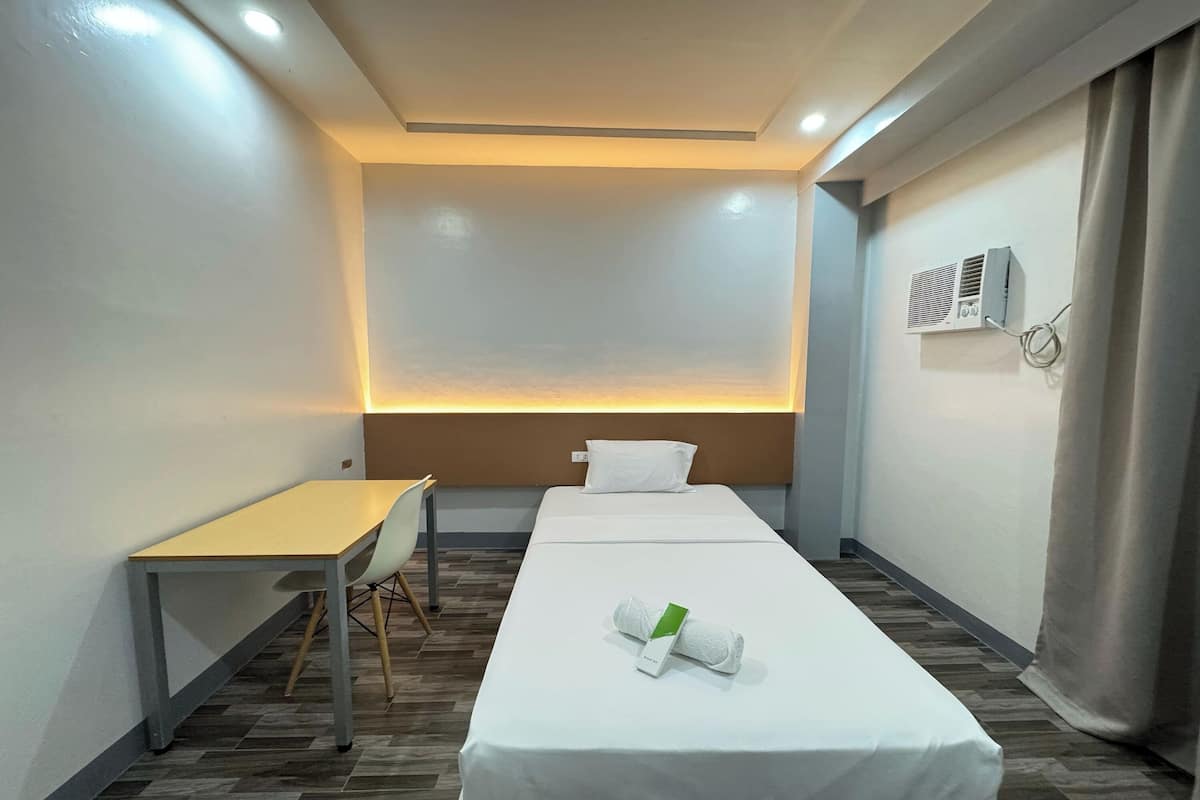 Ein Hotelzimmer mit einem Einzelbett, einem kleinen Schreibtisch, einem Sessel und einer Wand-Klimaanlage.