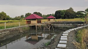 A rural setting with two wooden structures with red roofs, a water body, and a stone pathway. - Tres Marias Farmhouse with Pool (Cuyapo)