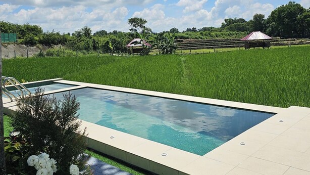 A rectangular swimming pool with a tiled deck, surrounded by a grassy area and a backdrop of a lush green field and trees. - Tres Marias Farmhouse with Pool (Cuyapo)