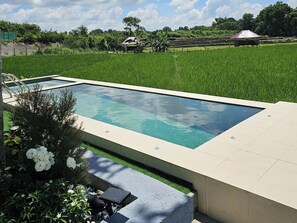 A rectangular swimming pool with a tiled deck, surrounded by a grassy area and a backdrop of a lush green field and trees. - Tres Marias Farmhouse with Pool (Cuyapo)