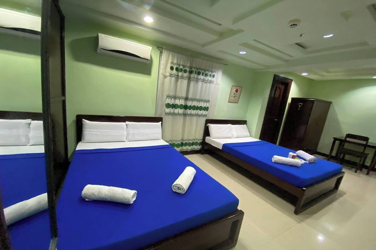 A hotel room with three single beds, each with a blue mattress cover and white pillows. There is an air conditioning unit mounted on the wall above the beds.