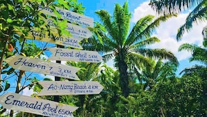 Ein Wegweiser mit mehreren Hinweisschildern, die zu verschiedenen Sehenswürdigkeiten wie einem Resort, einem Strand und Naturgebieten zeigen, umgeben von üppiger Vegetation.