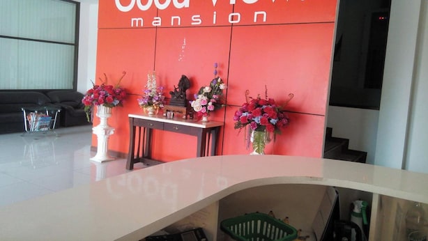 A reception area with a red wall displaying the text "Good View. mansion". There are three flower arrangements on the wall, a reception desk, and a sofa in the background. - Good View Residence (Lampang)