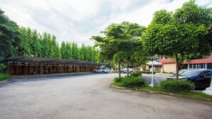 A parking lot with a covered area, trees, and buildings in the background. - Good View Residence (Lampang)