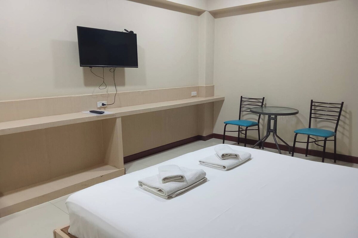 A hotel room with a bed, a TV mounted on the wall, a small table with chairs, and a shelf with a remote control.