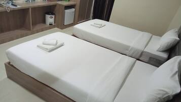 A hotel room with two single beds, a wooden nightstand, a small refrigerator, and a desk with a telephone and a green bag.