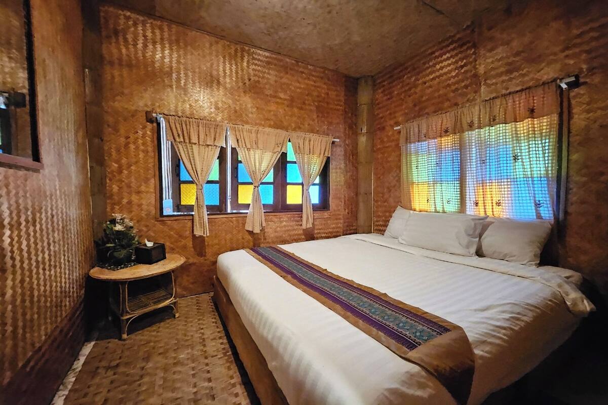 Un dormitorio con una cama grande, dos ventanas con cortinas, una mesita con una planta y un papel tapiz estampado.