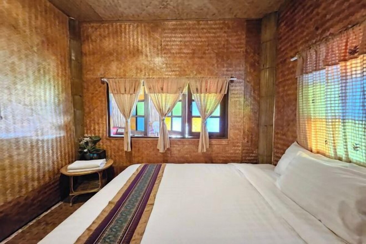 Een kamer met een groot bed, een klein tafeltje met een plant en een raam met gordijnen.