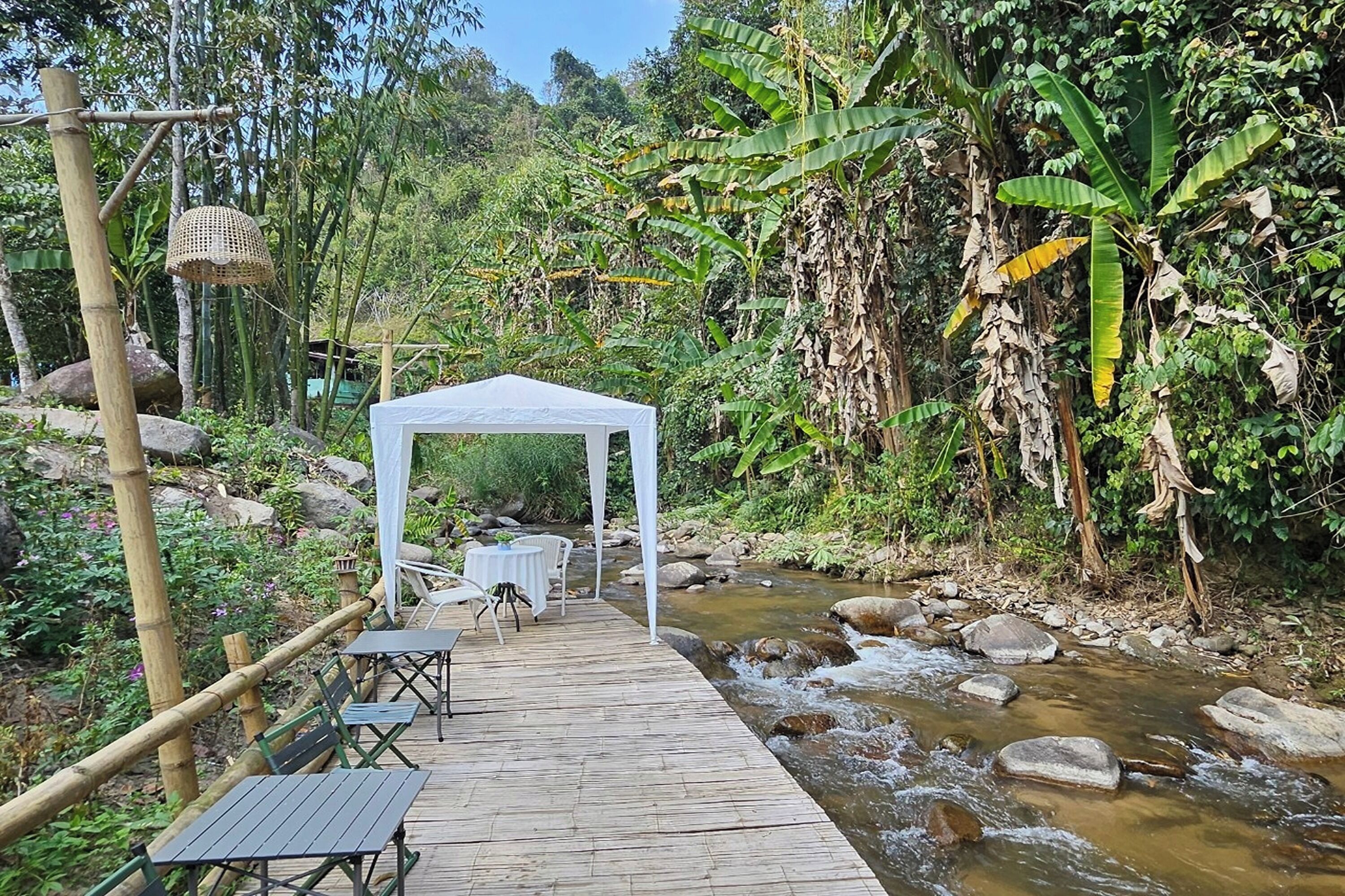 A wooden deck with tables and chairs, a white canopy, and a stream.
