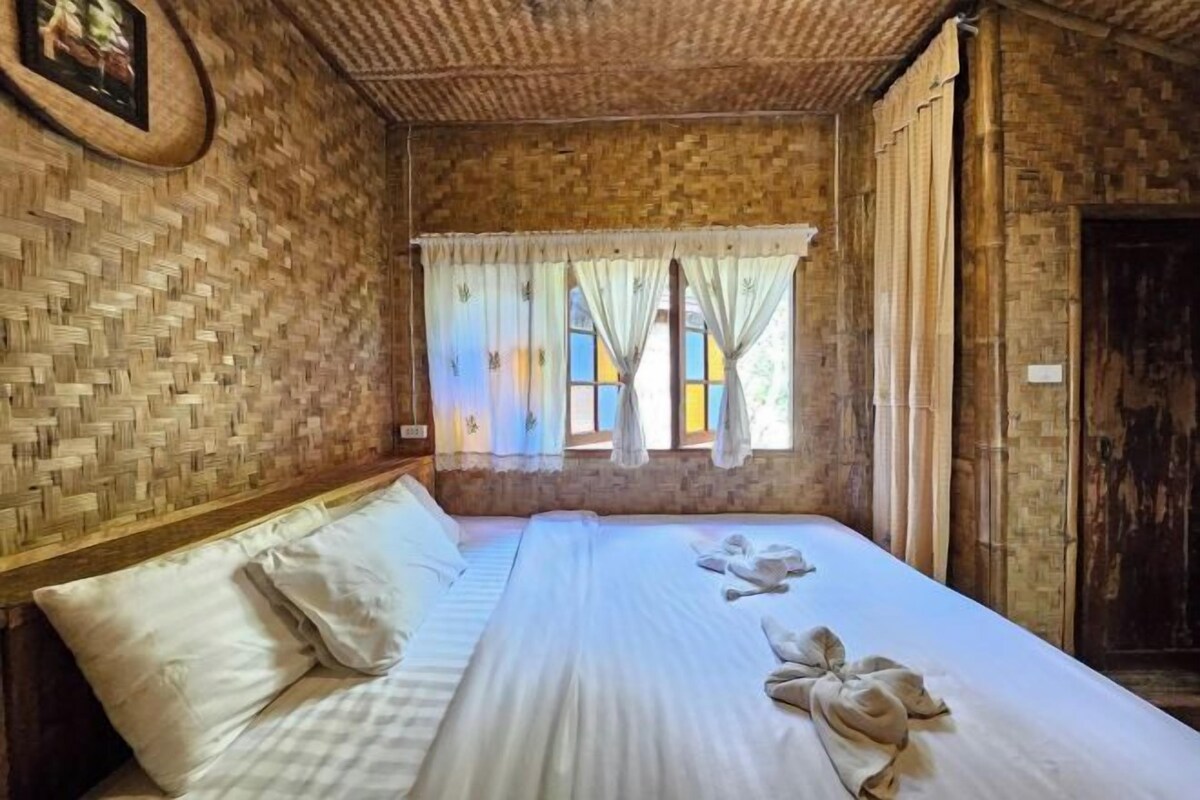 Une pièce dont le mur est décoré d’un motif tissé, avec un lit recouvert de draps blancs et une fenêtre ornée de rideaux légers.
