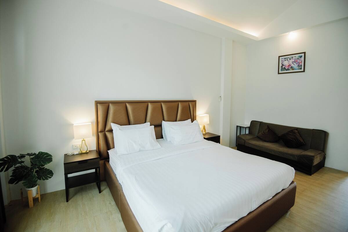 A bedroom with a large bed, a bedside table with a lamp, a small table with a plant, and a couch.