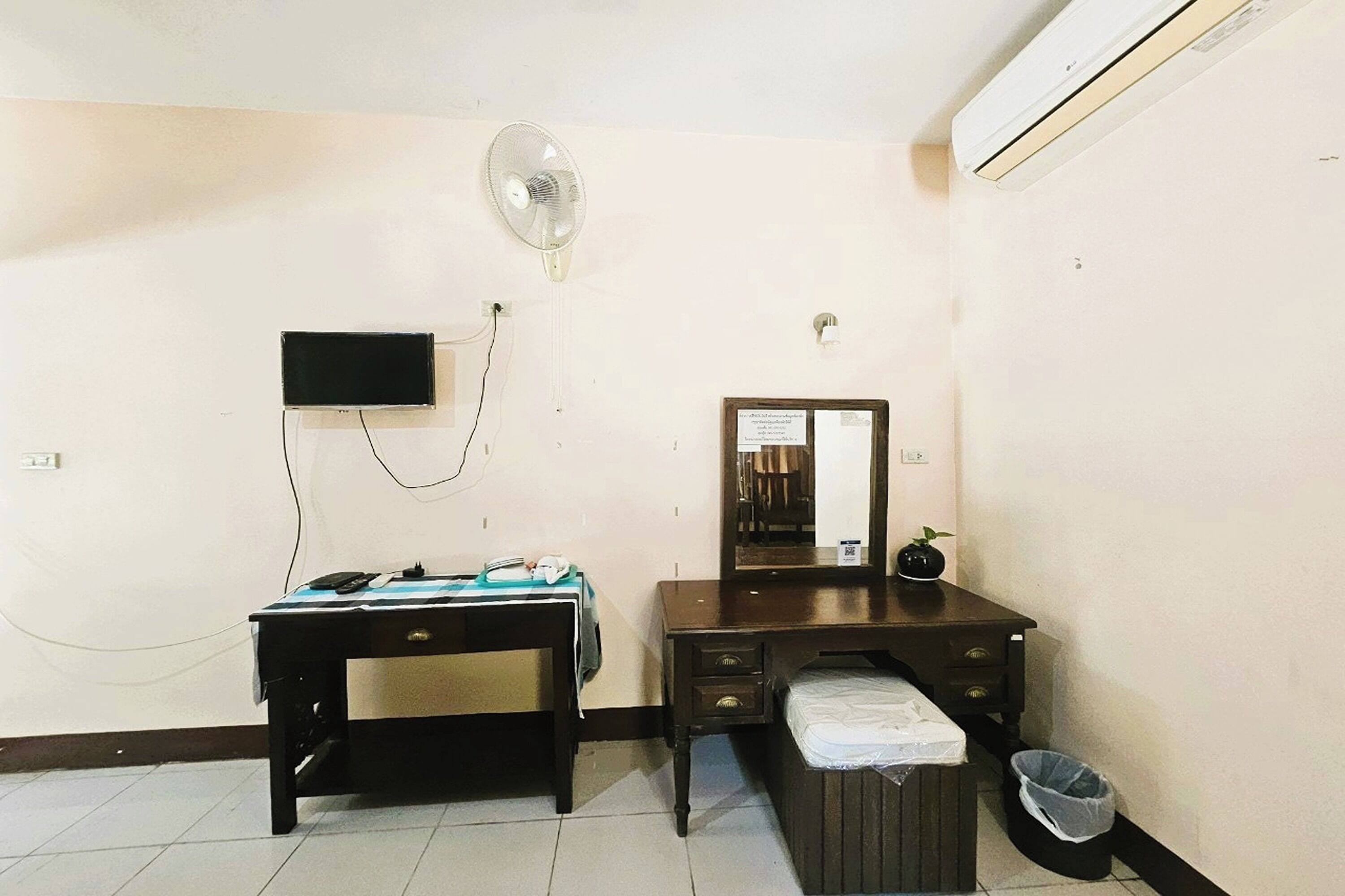 A small, clean room with a desk, a chair, a bed, a mirror, a fan, and a trash bin.