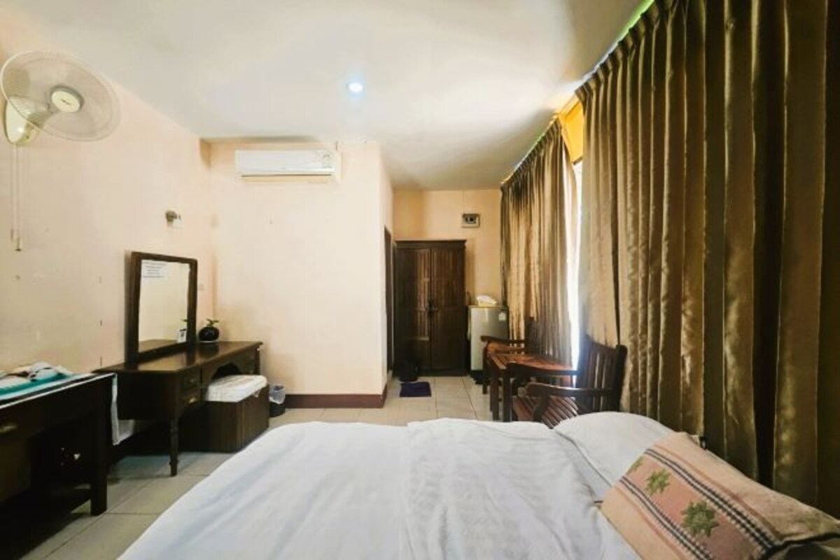 Habitación de hotel con cama, escritorio, silla, espejo, ventilador y ventana con cortinas.