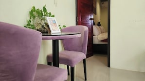 Una habitación con una pequeña mesa y dos sillas moradas, una planta sobre la mesa y una cama al fondo.