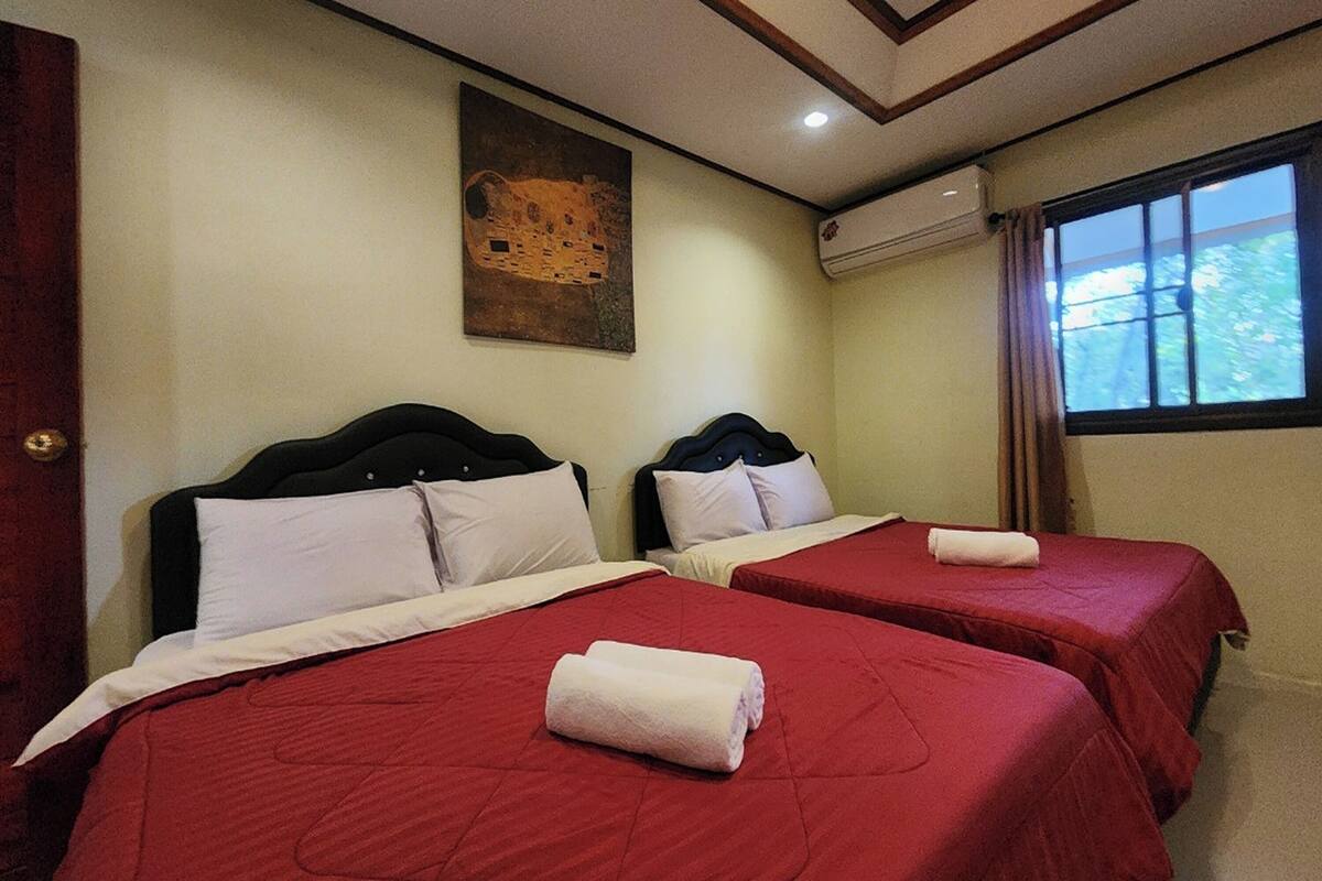 Habitación de hotel con dos camas, cada una con ropa de cama roja y almohadas blancas. Hay un cuadro en la pared y un equipo de aire acondicionado sobre la ventana.