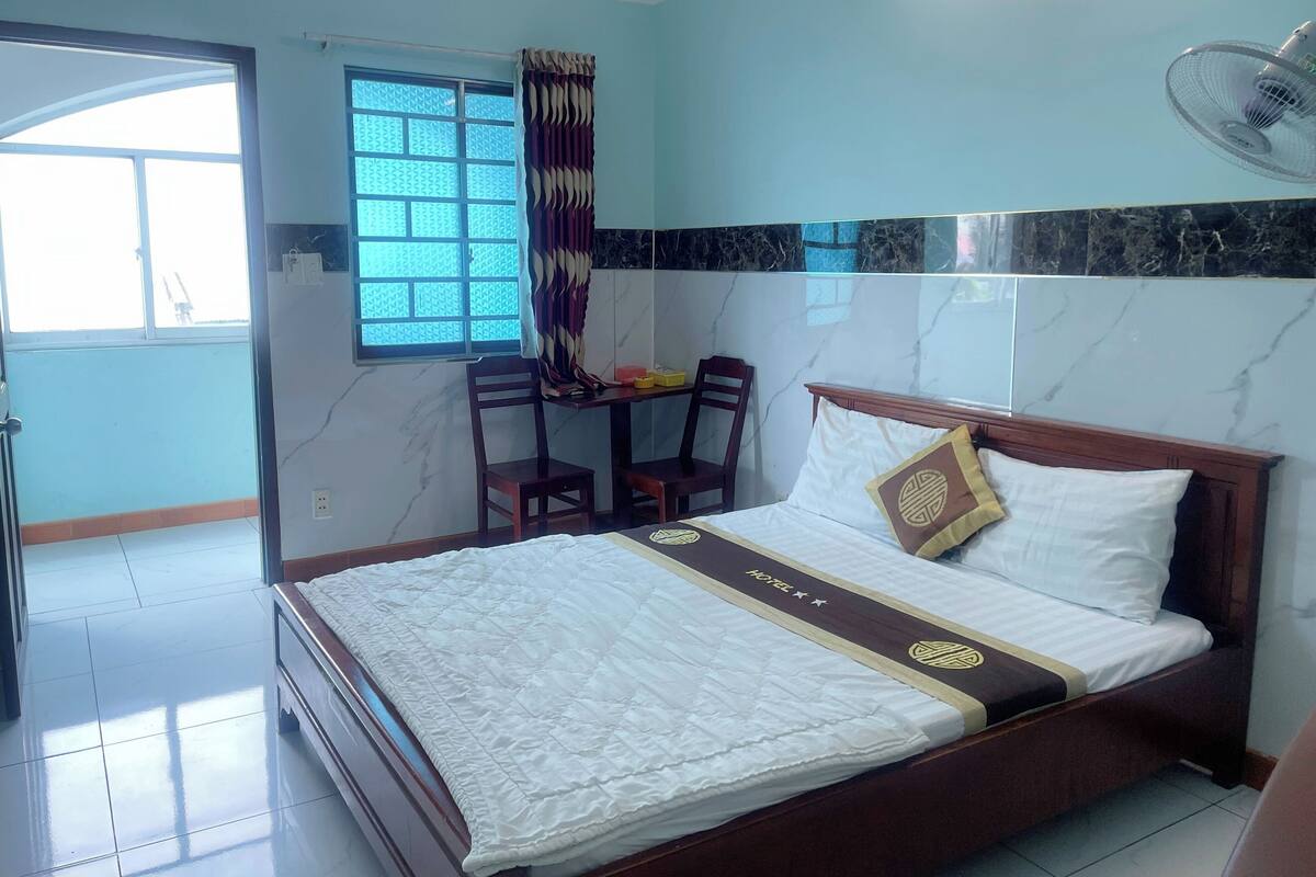 A hotel room with a bed, a desk with chairs, a window with curtains, and a wall-mounted fan.