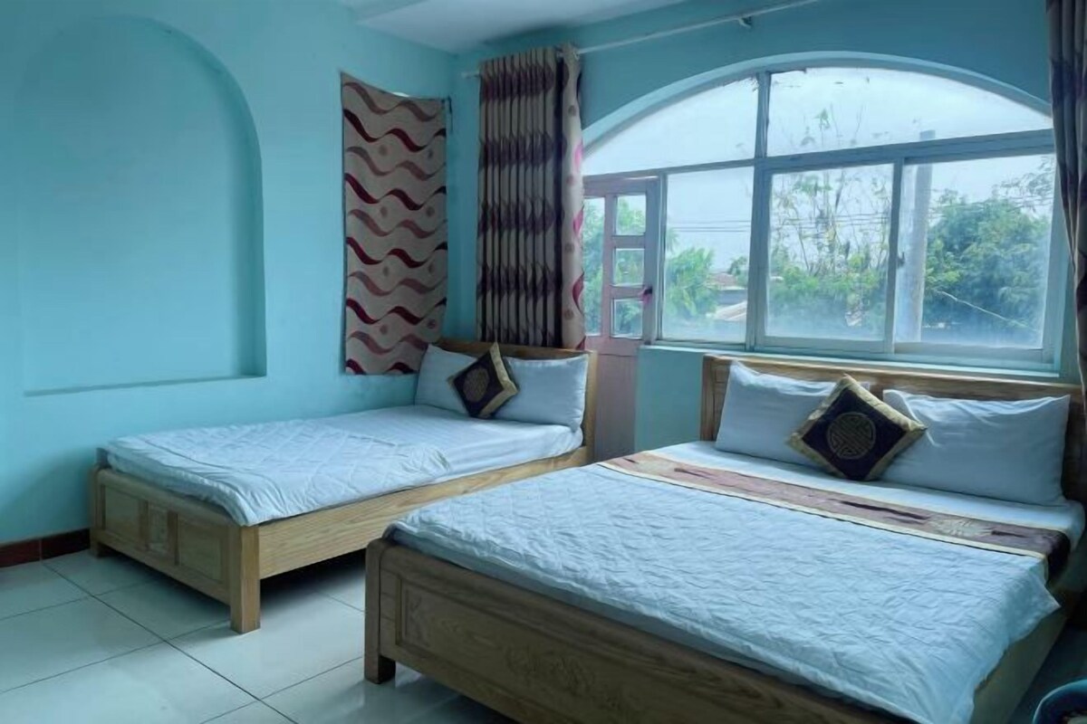 Quarto com duas camas de madeira, janela com vista para vegetação e cortina estampada.