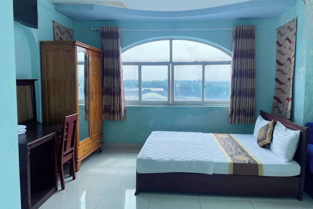 A bedroom with a bed, a wooden wardrobe, a desk, and a large window with curtains.