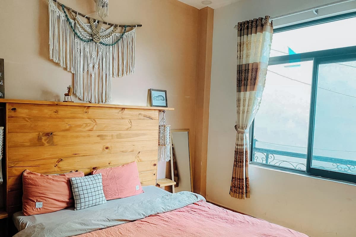 A bedroom with a wooden headboard, a bed with pink bedding, a macrame wall hanging, and a window with patterned curtains.