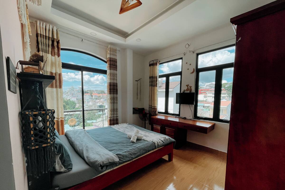 A bedroom with a bed, a desk, and large windows offering a city view.