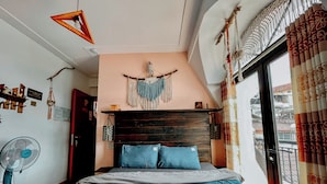 Une chambre avec un lit, un ventilateur, une décoration murale en macramé et une vue sur l’extérieur.