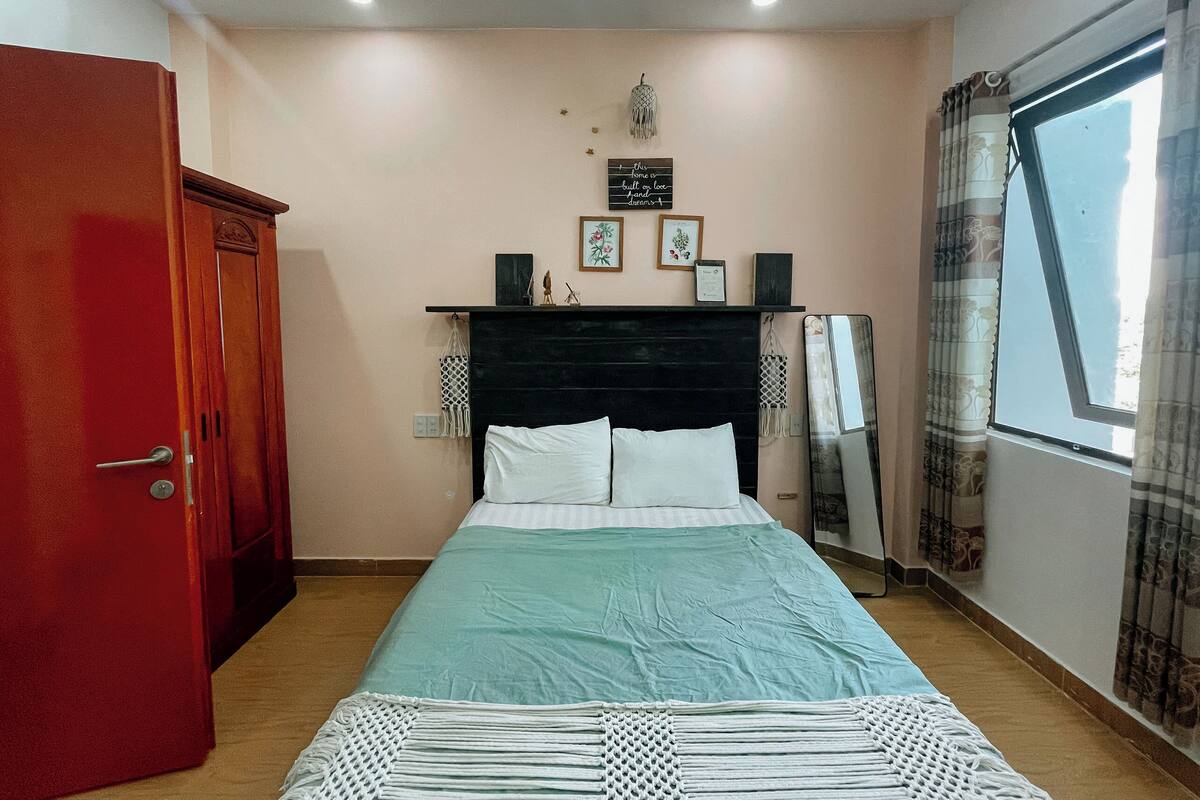 A bedroom with a bed, a wardrobe, a shelf with decorative items, a window with curtains, and a wall-mounted mirror.
